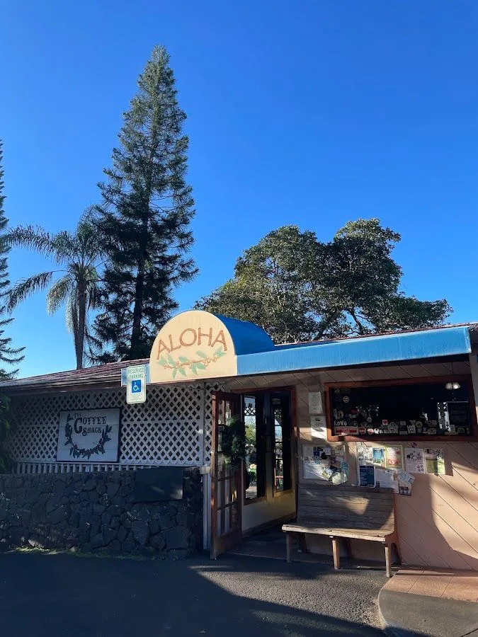 The Coffee Shack exterior with an ALOHA awning, lattice wall, wooden bench, and tall trees.