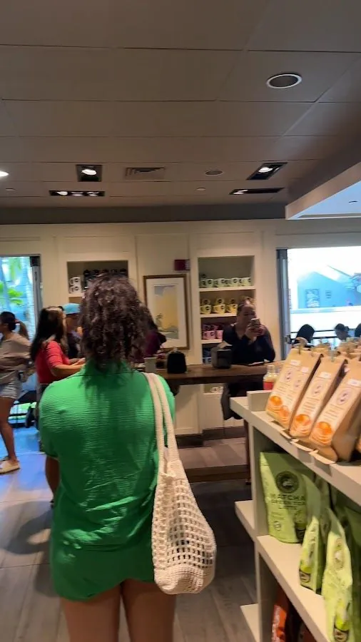 A person in green with a white woven bag faces a shop counter, other people stand nearby, and packaged bags sit on a shelf.