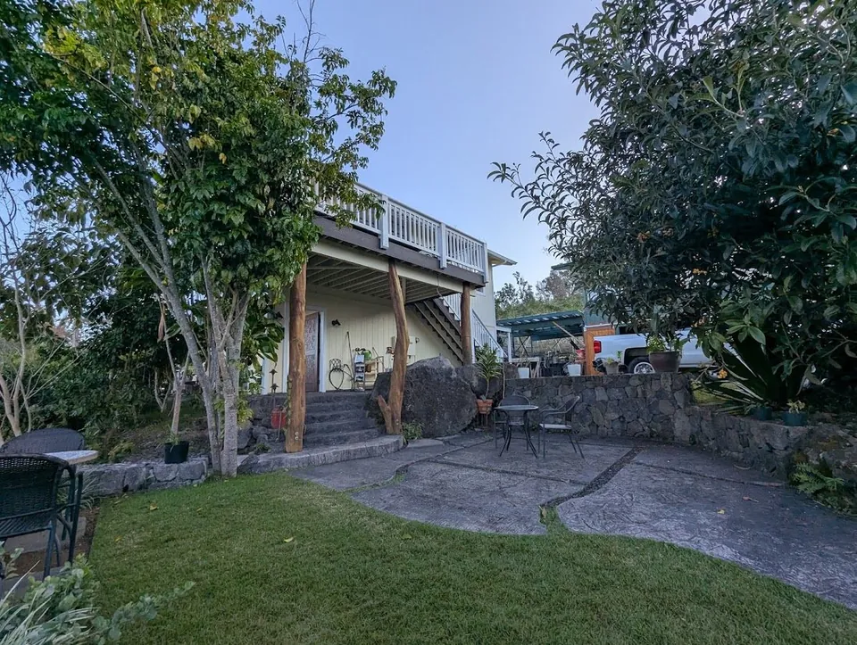 Pale house with a raised white-railed deck, lawn, patio chairs, trees, a stone wall, and a white truck.