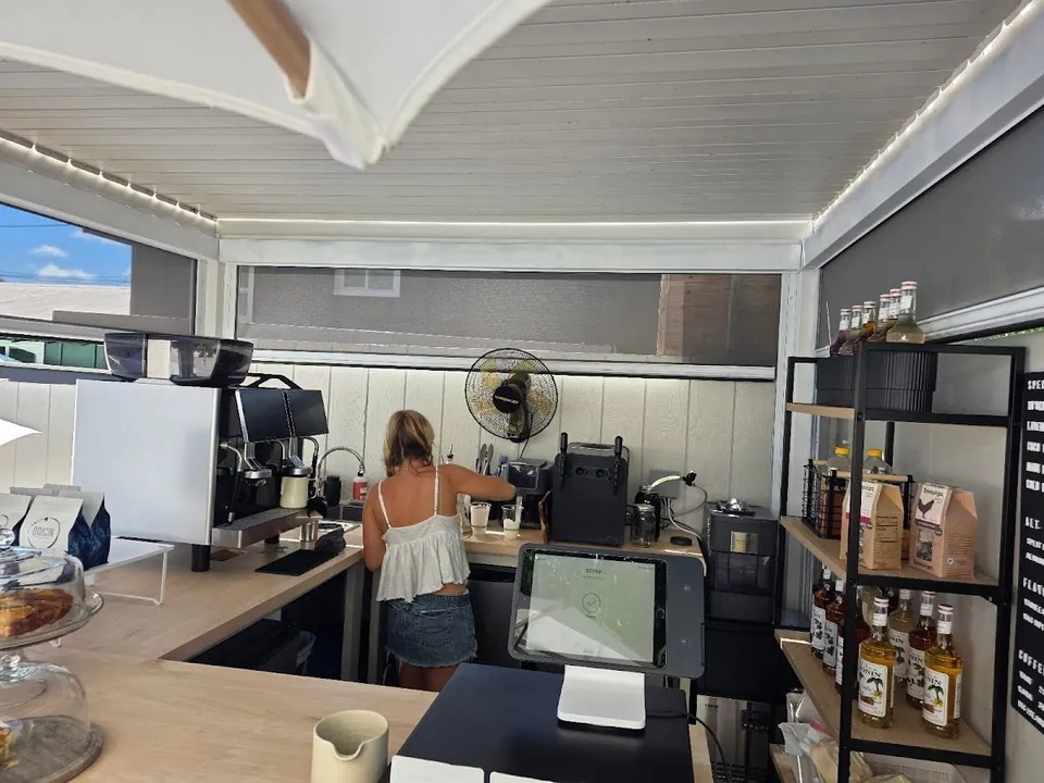 A person works at an espresso counter inside a bright café, with a POS tablet, shelves of bottles, and a cropped menu board.