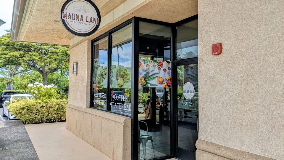 Beige stucco storefront of Mauna Lani Coffee Co. with a round hanging sign, lettered glass windows, and a glass door.