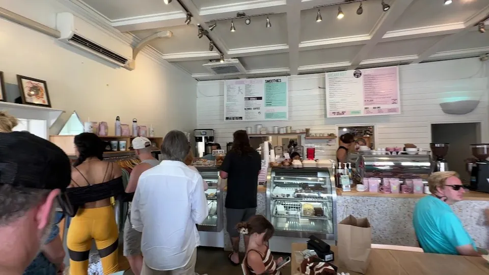 People wait at a cafe service counter under wall menu boards, next to a glass display case and coffee grinders.