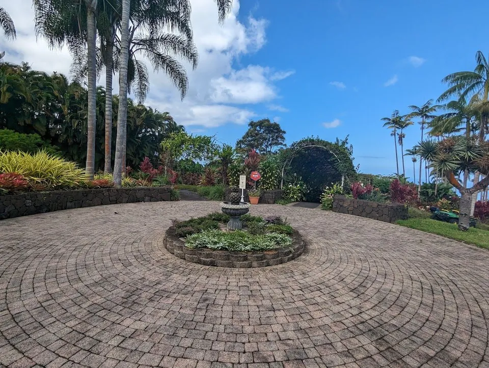 Circular paver courtyard with a stone fountain bed, palms, lava-rock walls, and tropical plants under a blue sky.