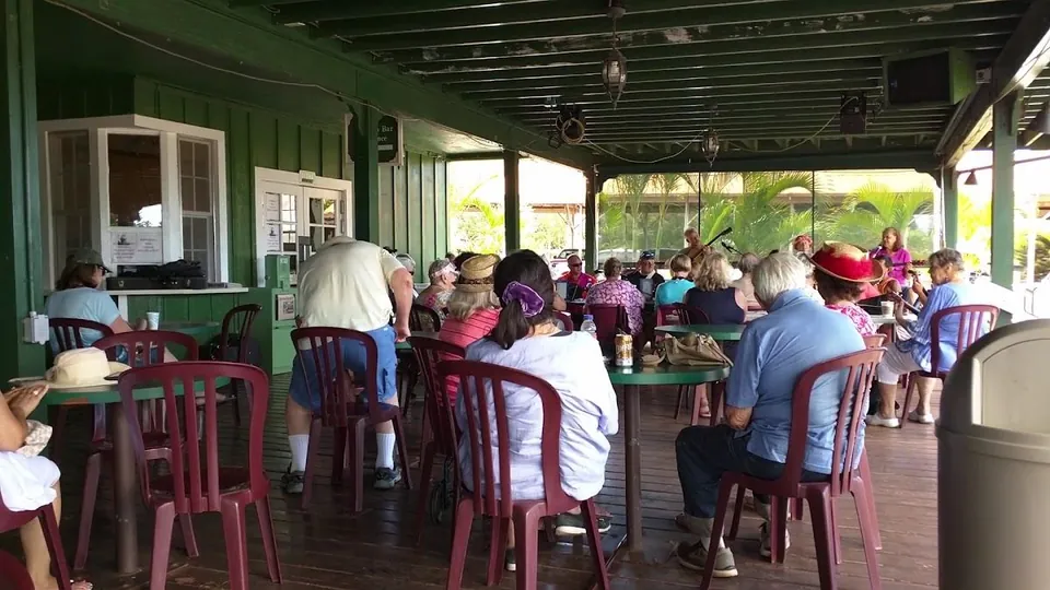 Covered green dining porch with people seated at round tables, maroon chairs, and a musician near the open side.