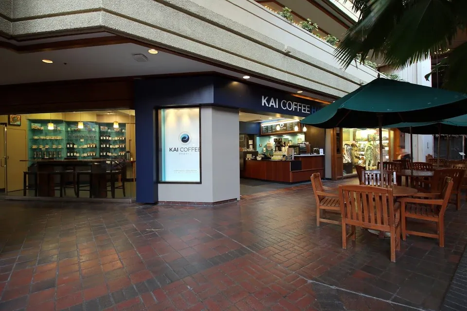 Kai Coffee storefront on a brick walkway, with window displays and wooden tables under green umbrellas.