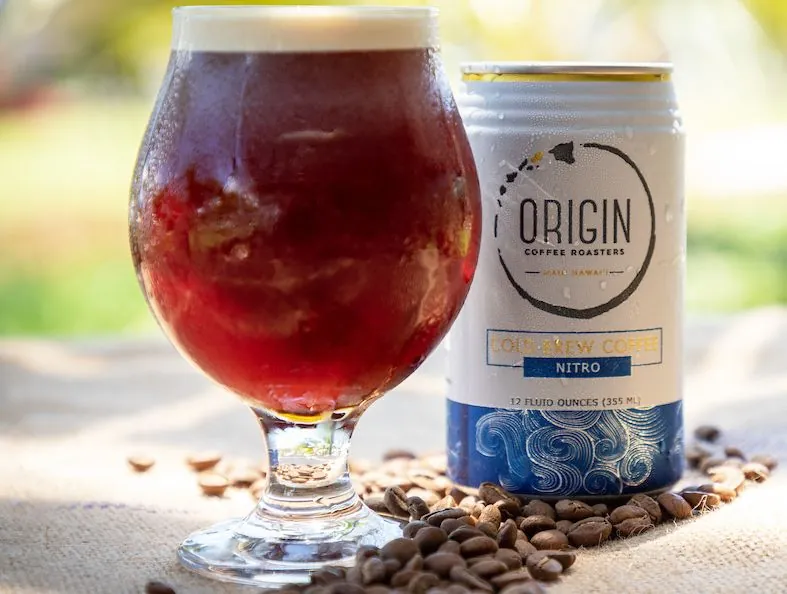 A stemmed glass of dark cold brew with foam beside a white Origin Coffee Roasters nitro can and coffee beans.