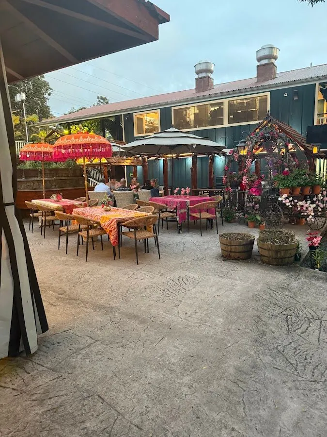 Diners sit at cloth-covered tables on a stamped-concrete patio with umbrellas, flowers, and a dark green building.