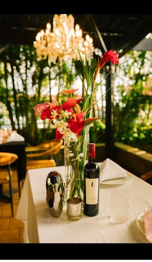 A white-clothed dining table holds red and white flowers, a dark wine bottle, and glasses beneath a chandelier.