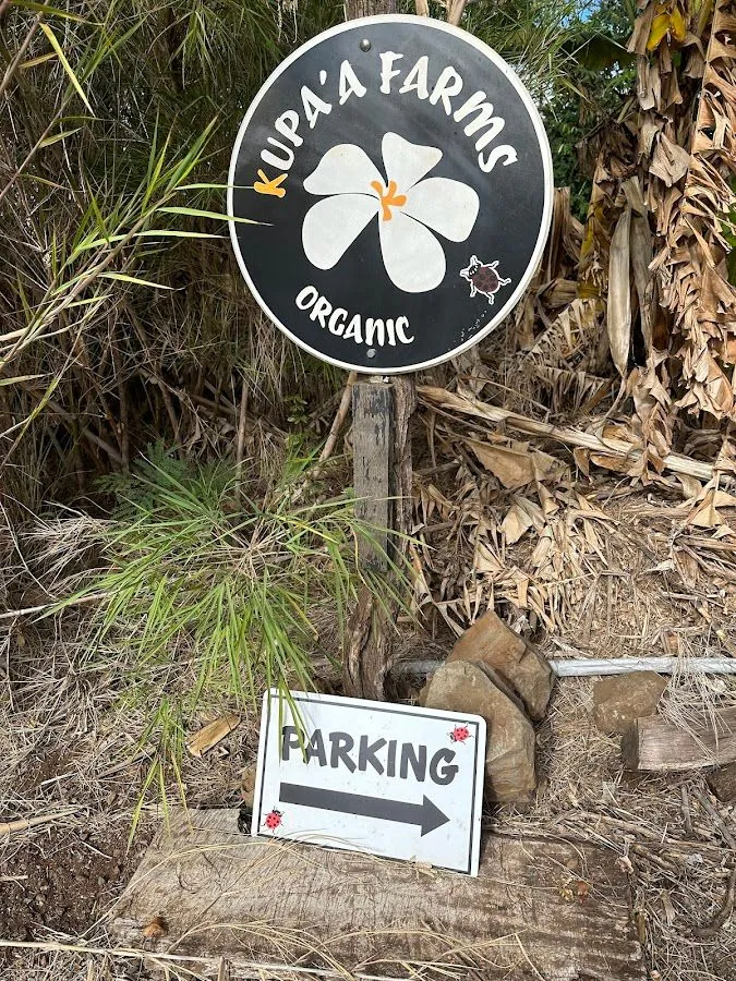A round Kupa'a Farms Organic sign stands above a Parking sign with a right arrow, beside dry leaves and rocks.