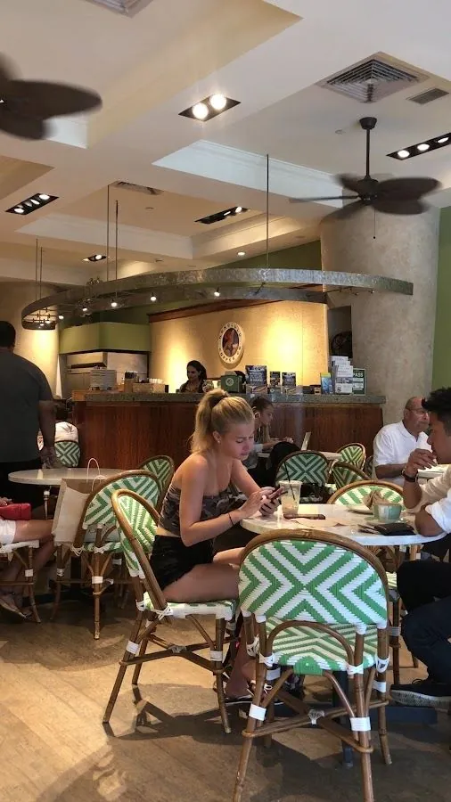 A person looks at a phone at a cafe table among green-and-white chairs, with a service counter and ceiling fans behind.