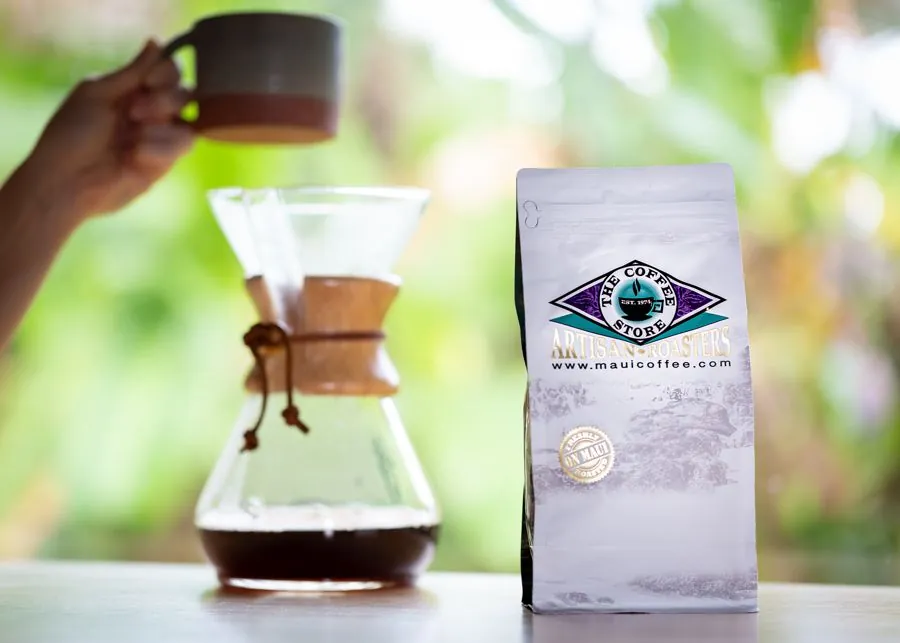 A hand holds a mug above a glass pour-over carafe of coffee beside a white The Coffee Store bag.