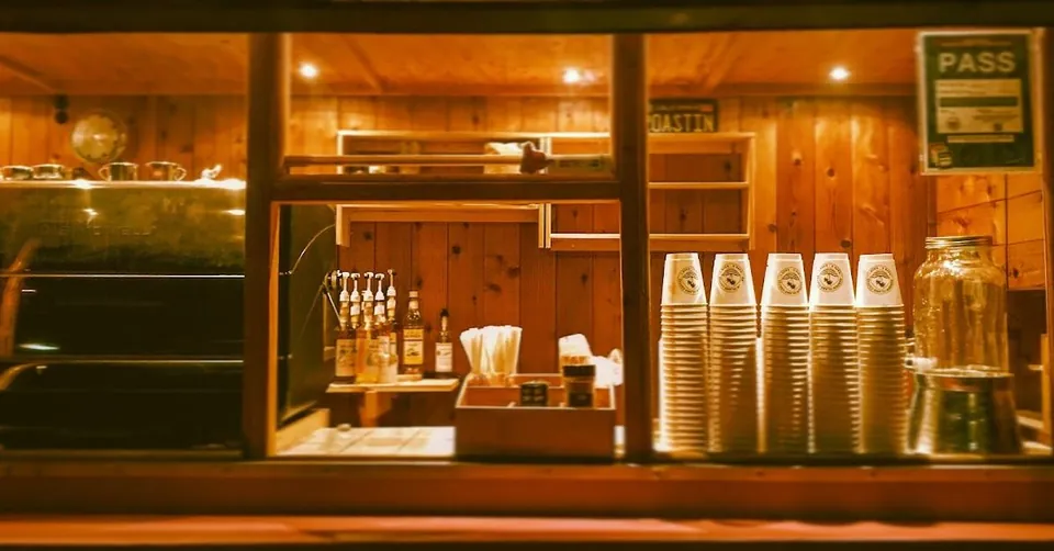 Wood-paneled coffee service counter with an espresso machine, stacked paper cups, pump bottles, and a PASS sign.