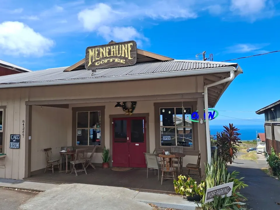 Beige coffee shop with a metal roof, porch tables, red doors, and an ocean view under a blue sky.