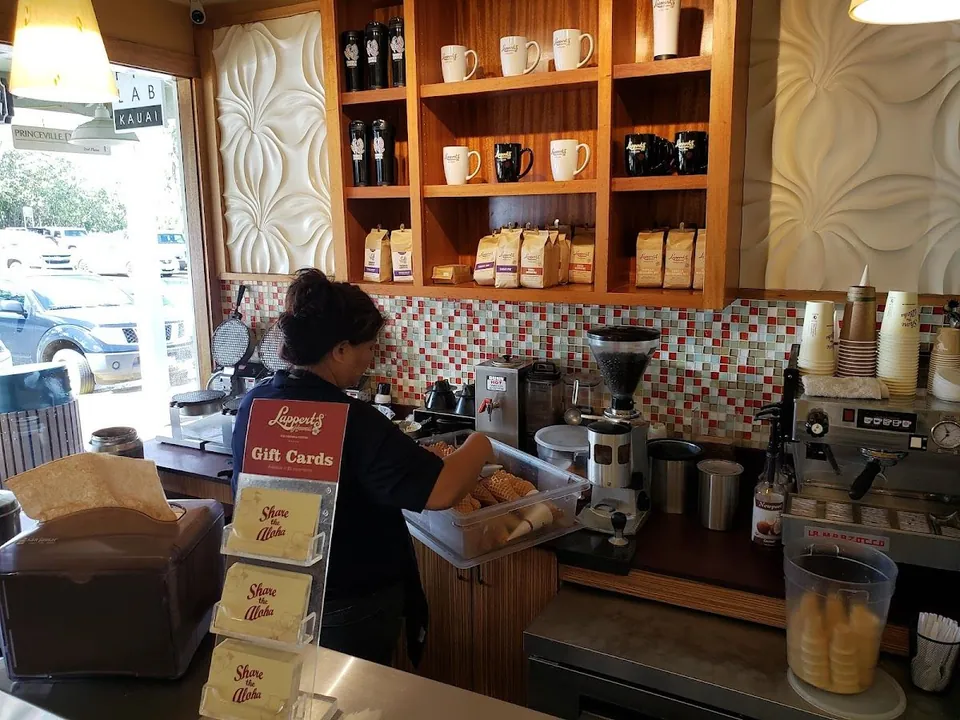 A worker reaches into a bin of cones behind a counter with a coffee grinder, espresso machine, and shelves.