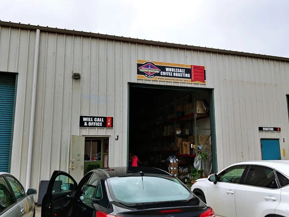 Person in a pink shirt stands in the open bay of a Maui Oma Coffee Roasting warehouse, with cars parked in front.