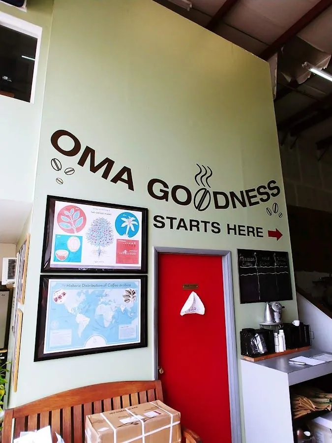 Pale green wall lettering reads OMA GOODNESS STARTS HERE above a red door, coffee posters, and a bench.