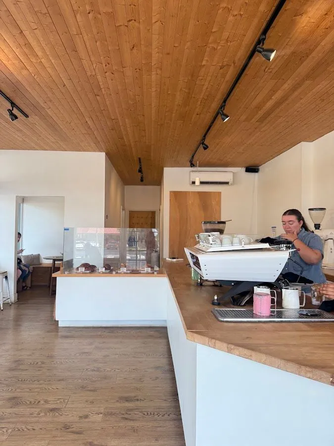 Cafe service counter under a wood ceiling, with a white espresso machine, a glass display case, and a worker behind the bar.
