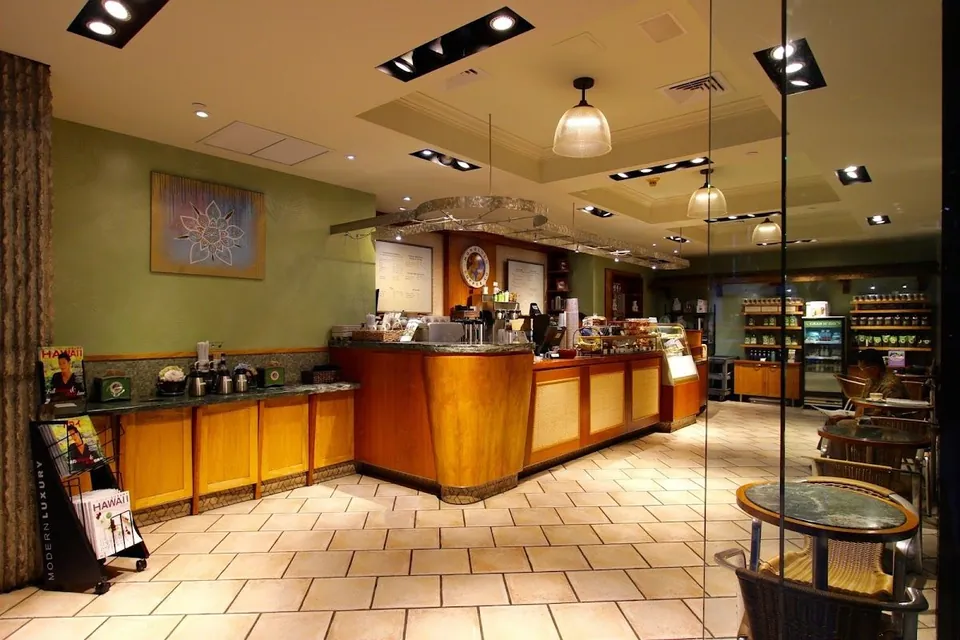 Wooden cafe counter with menu boards and a display case; tables and one seated person by a glass wall.