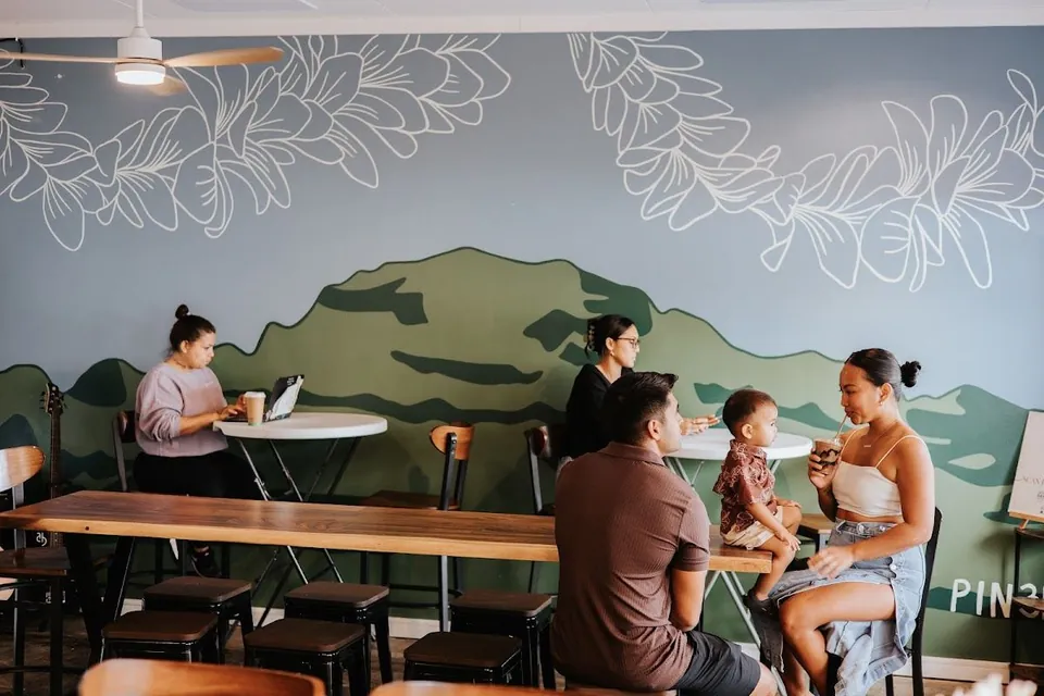 People sit at tables in a cafe with a floral mural; one uses a laptop and one holds a clear cup.