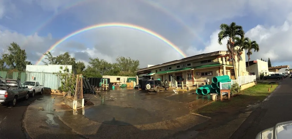 A double rainbow arches over wet pavement, low buildings, parked trucks, and stacked green pipes.