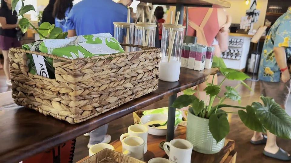 Woven basket of green patterned bags, glass tumblers with straws, plants, and mugs on shop shelves, people standing nearby.