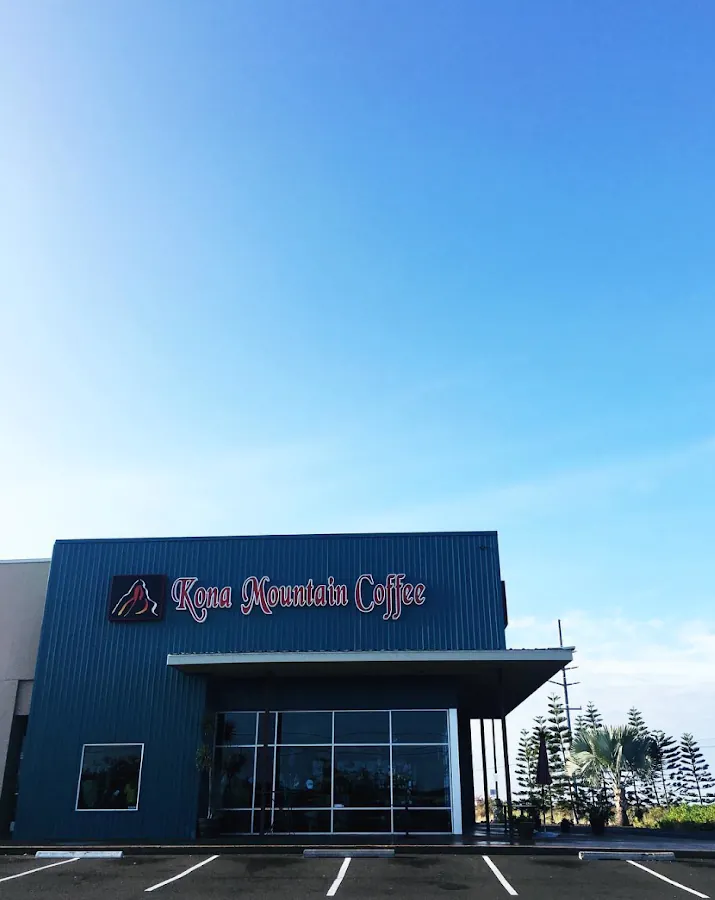 Blue metal Kona Mountain Coffee storefront with a glass entrance, parking spaces, and a clear sky.