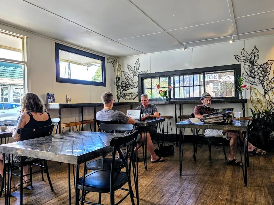 People sit at metal tables in a bright cafe; one uses a laptop, with wood floors and botanical wall drawings.