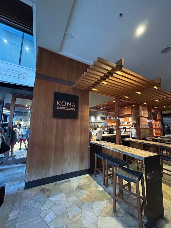Cafe interior with a Kona Coffee Purveyors wall sign, a wood bar and stools, and people near the counter and door.