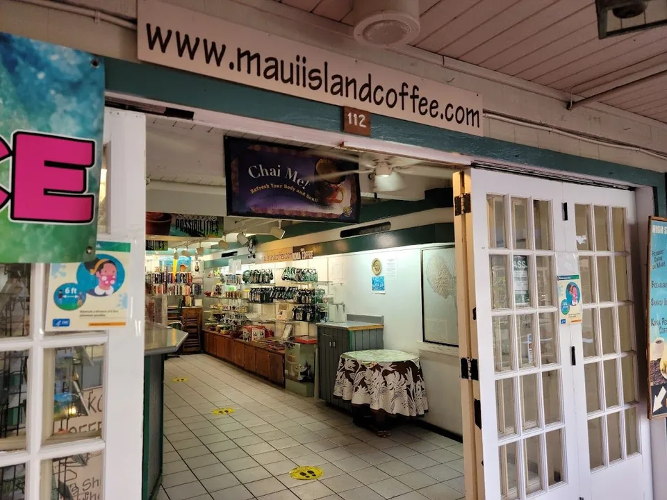 Open French doors under a www.mauiislandcoffee.com sign look into a shop with shelves, a chai banner, and a cloth-covered table.