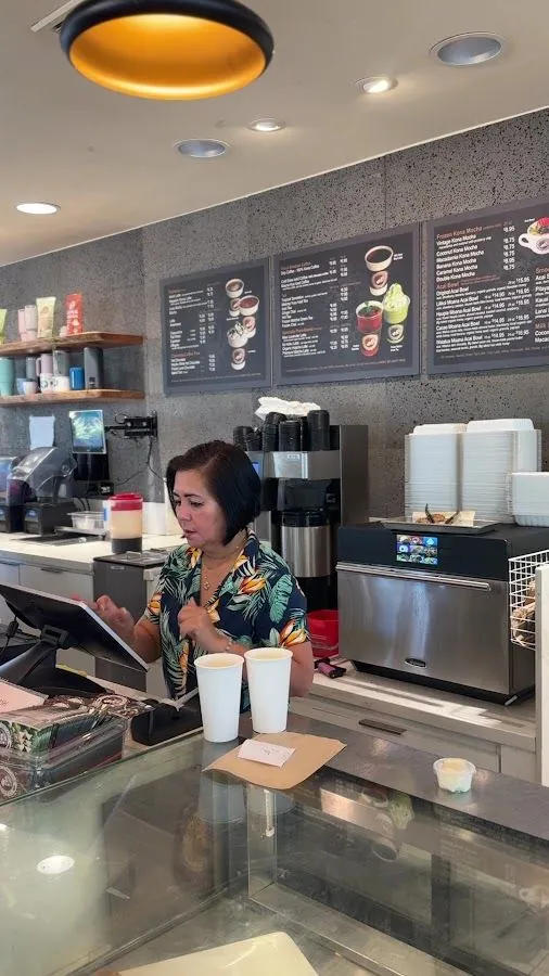 A worker at a cafe counter uses a touchscreen register, with two paper cups and wall menu boards behind.