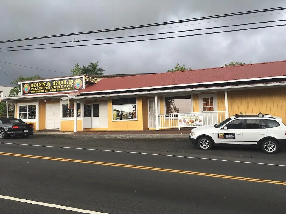 Yellow storefront with a red roof and a Kona Gold Trading Company sign beside a road under gray skies.
