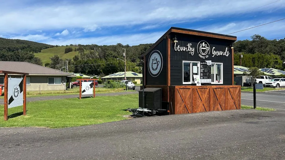 Black roadside stand labeled Country Grounds, with a wood base, on a lawn by a road and green hills.