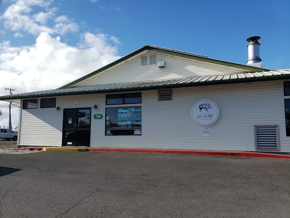 White single-story building with a metal roof, coffee window lettering, a round wall sign, and a parking lot.
