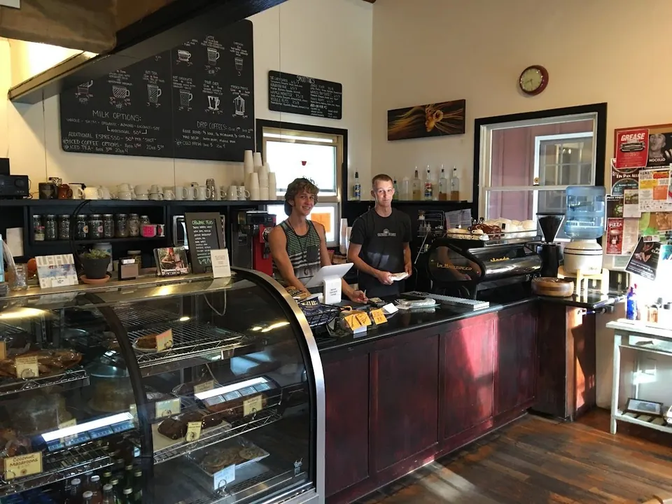 Coffee shop counter with two staff members, a pastry case, and chalkboard menus on the wall.